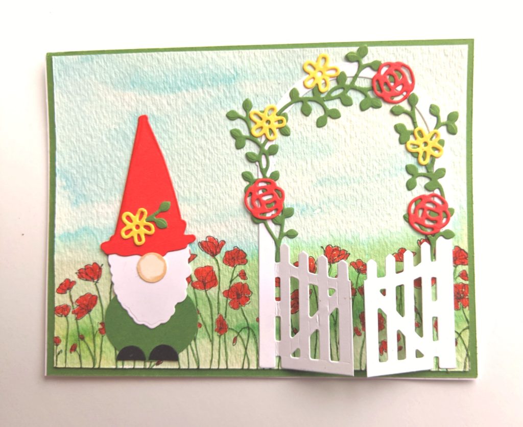 card with a paper gnome and poppies in the background and a white fence with a gate